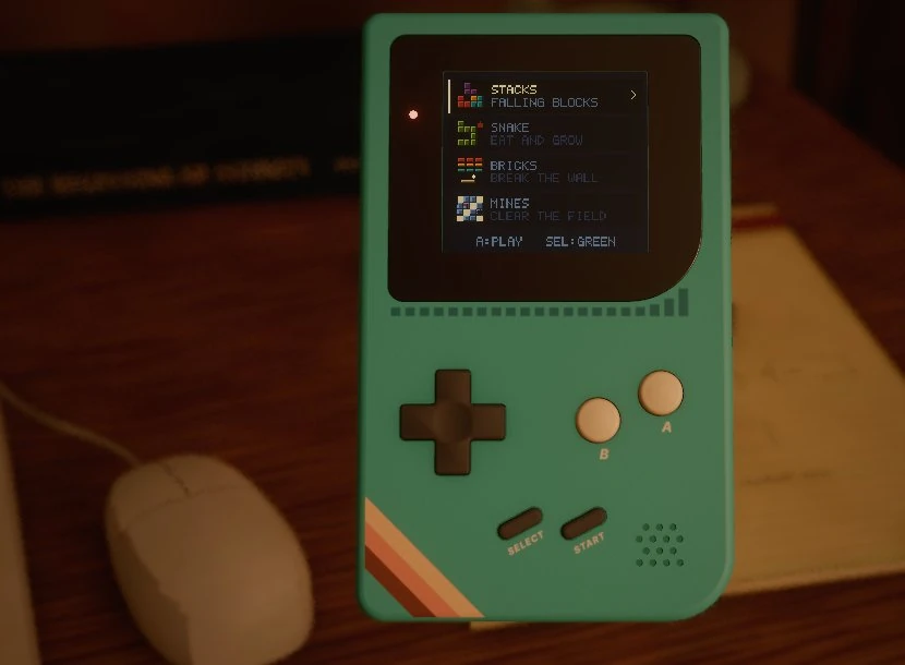 Close-up of the teal handheld on the desk next to the mouse, its screen showing a menu: Stacks, Snake, Bricks and Mines